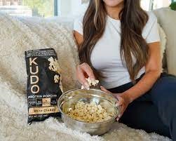 Female sitting on her sofa enjoying a Kudos healthy protein popcorn snack.